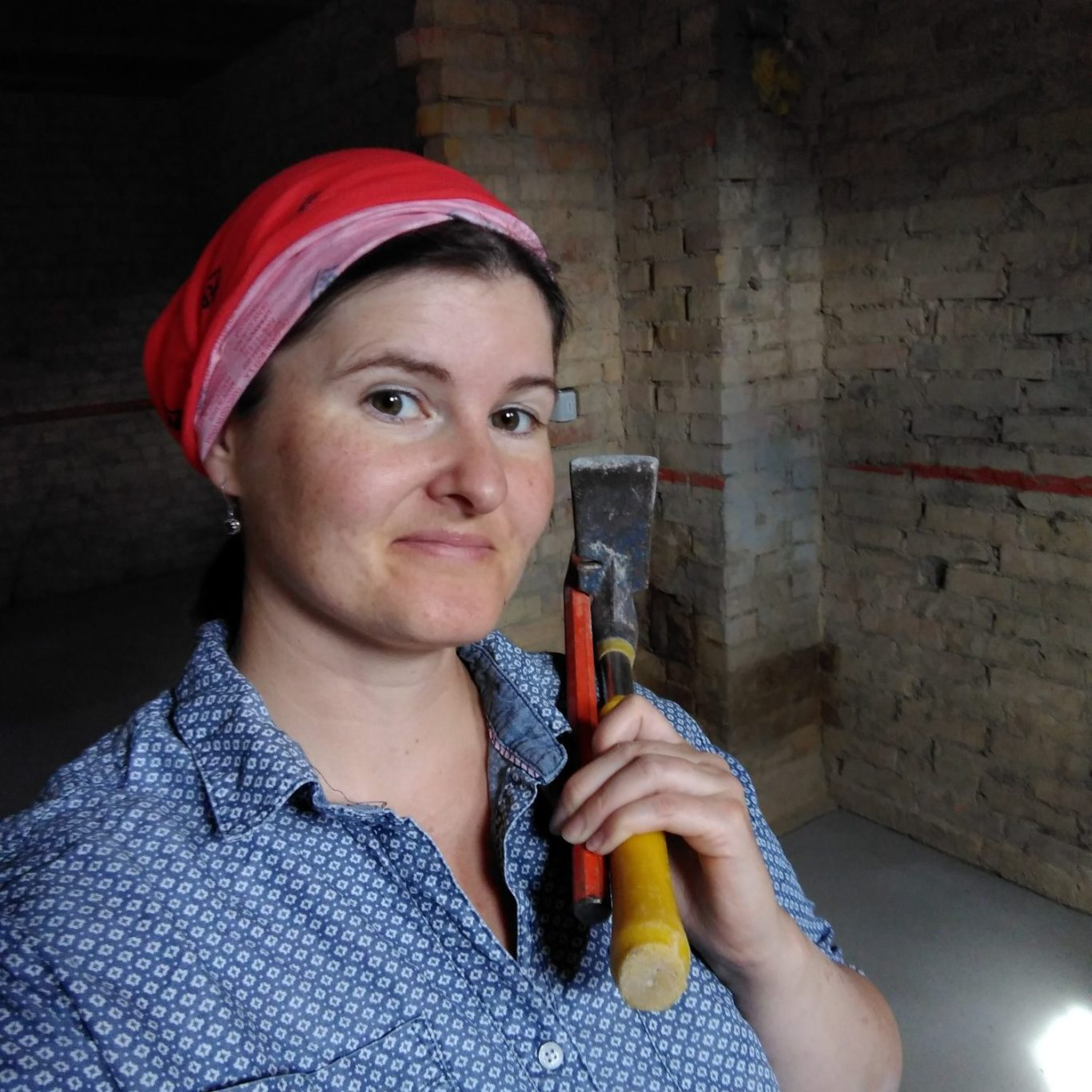 Bild zeigt Marie, von Studio Marie: Brünett mit rotem Kopftuch, Hammer und Stemmeisen in der Hand. Im Hintergrund eine rohe Steinmauer.